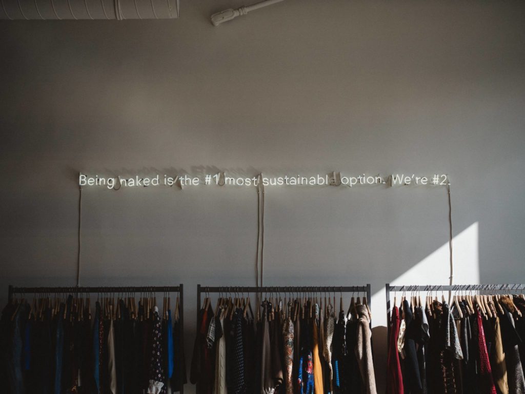 Blick in einen hippen Kleiderladen, in dem über den Kleiderständern eine Leuchtschrift die Aussage macht, dass Nacktheit die Nummer 1, der Shop aber die Nummer 2 in Sachen Nachhaltigkeit für Bekleidung sei.  