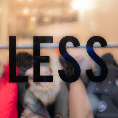 Das Wort "LESS" auf einer Schaufensterscheibe zeigt im Hintergrund Textilien, die auf einer Kleiderstange aufgehängt sind. Es ist ein Kommentar und Kritik am Konsum und der Verschwendung der Mode-Industrie. Photo by theblowup bei Unsplash