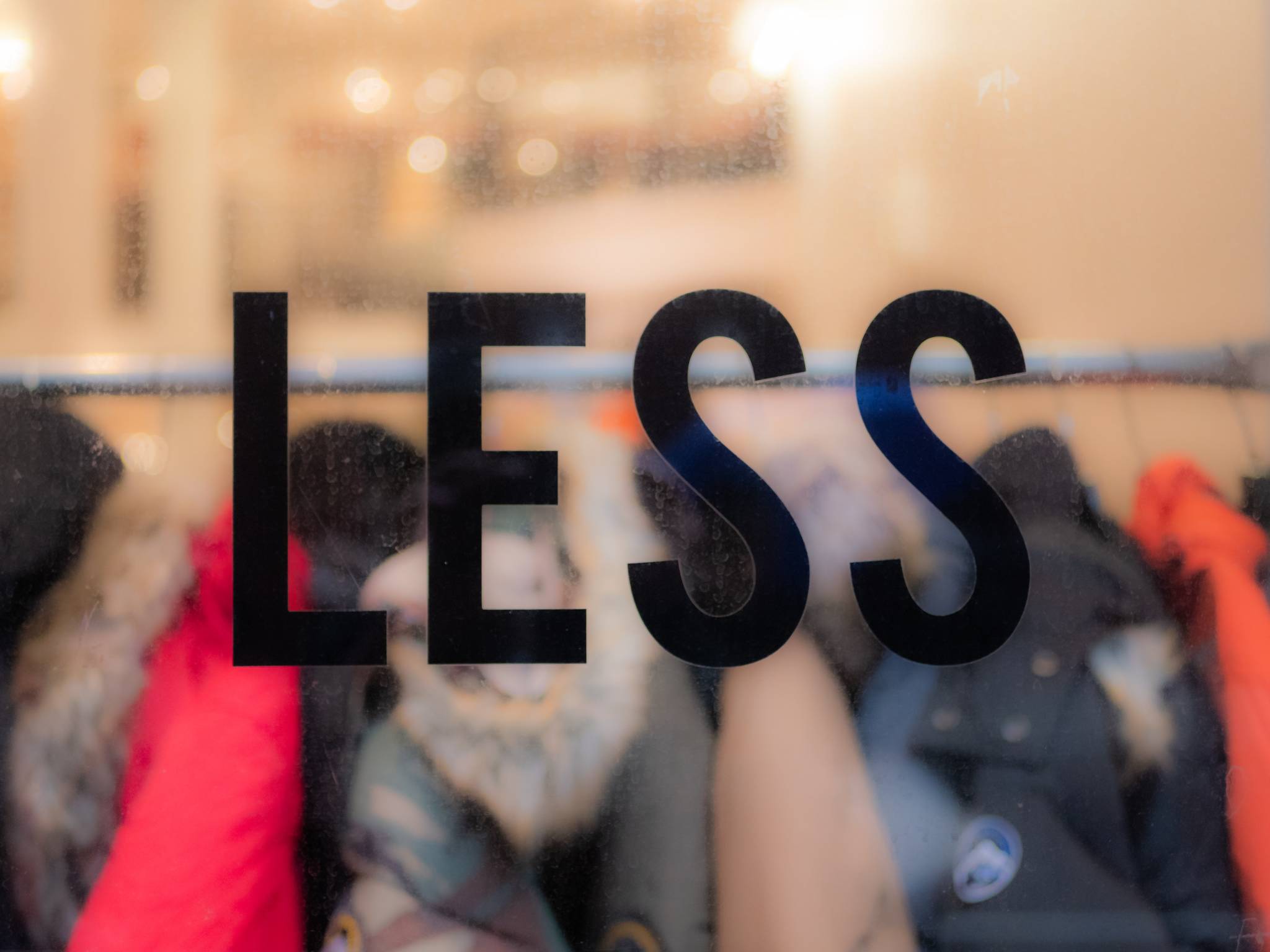 the-blowup-TDOQPhq631s-unsplash (2) Das Wort "LESS" auf einer Schaufensterscheibe zeigt im Hintergrund Textilien, die auf einer Kleiderstange aufgehängt sind. Es ist ein Kommentar und Kritik am Konsum und der Verschwendung der Mode-Industrie. Photo by theblowup bei Unsplash